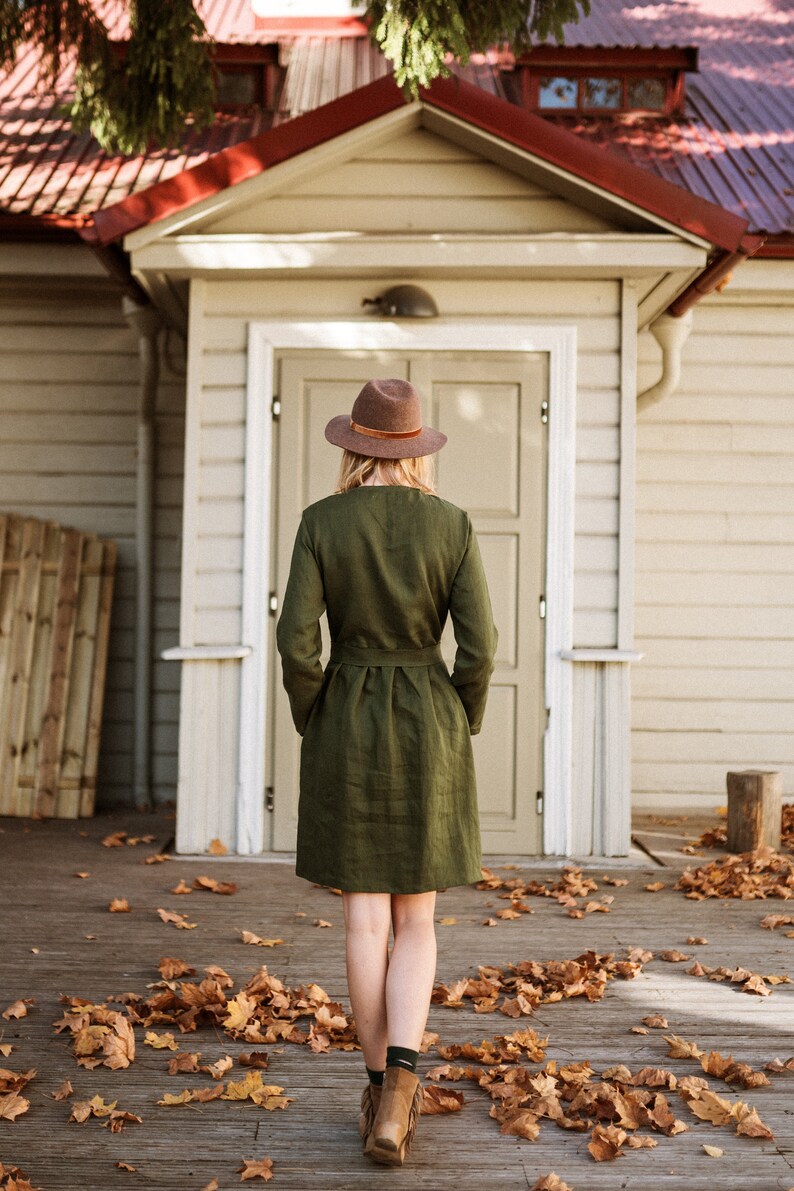 May include: A woman wearing a green linen dress and a brown hat stands in front of a white wooden building with a red roof.  She is walking on a wooden deck covered in fallen autumn leaves.