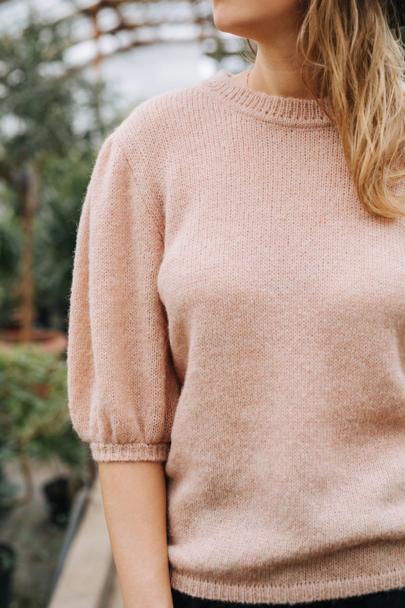 May include: A soft, pale pink knitted sweater with a round neckline and slightly puffed sleeves.