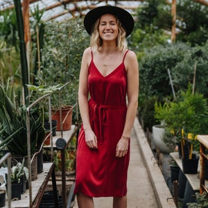 May include: A woman wearing a red dress and a black hat stands in a greenhouse. She is smiling and looking at the camera.
