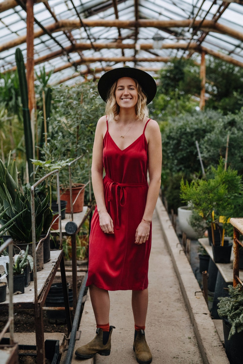 May include: A woman wearing a red dress and a black hat stands in a greenhouse. She is smiling and looking at the camera.