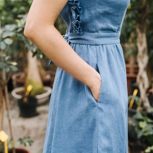 Puede incluir: Una mujer lleva un vestido de lino azul claro con un detalle de volantes en la espalda. El vestido tiene un corpi&ntilde;o ajustado y una falda fluida. La mujer est&aacute; de pie en un entorno de jard&iacute;n.