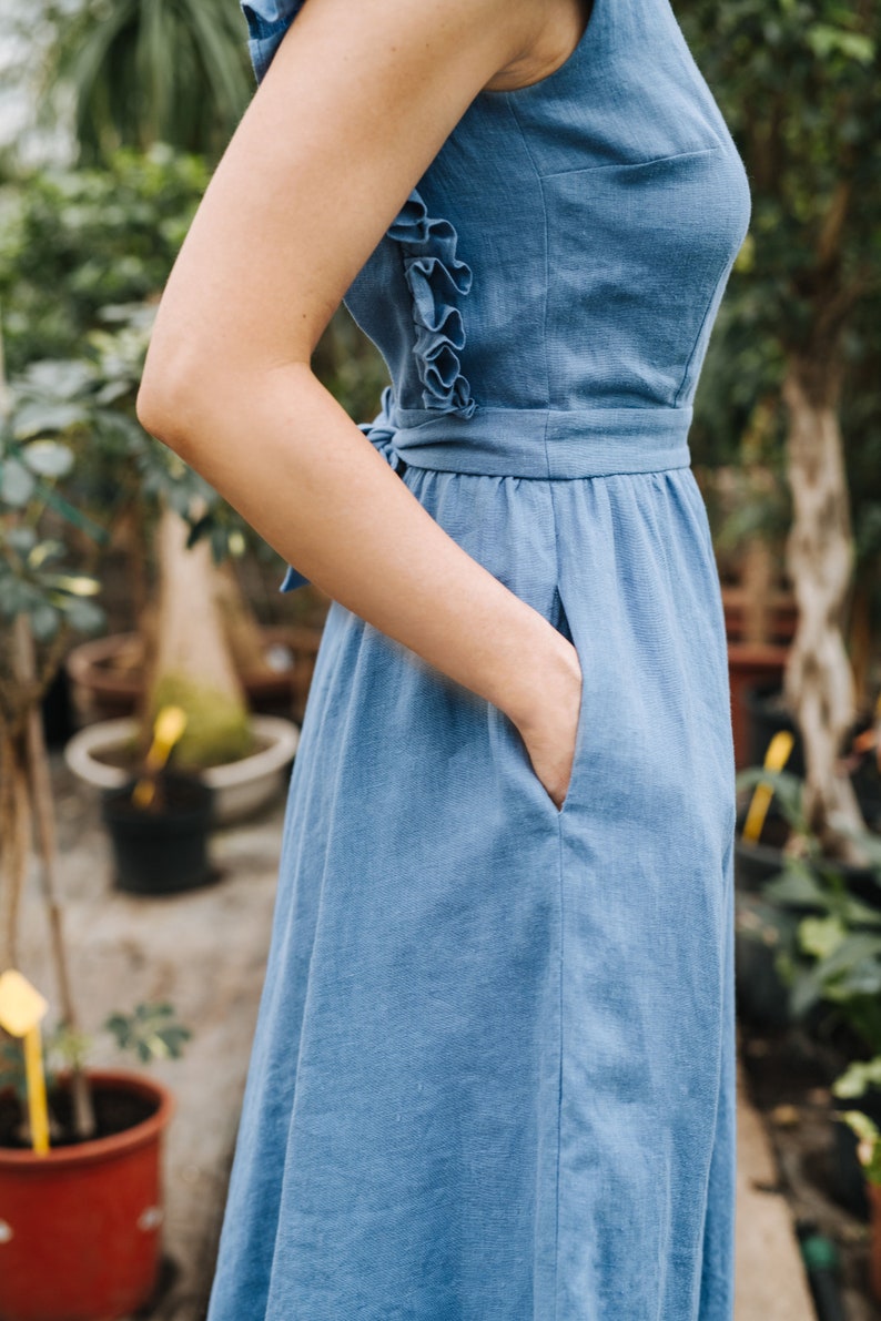 Puede incluir: Una mujer lleva un vestido de lino azul claro con un detalle de volantes en la espalda. El vestido tiene un corpi&ntilde;o ajustado y una falda fluida. La mujer est&aacute; de pie en un entorno de jard&iacute;n.
