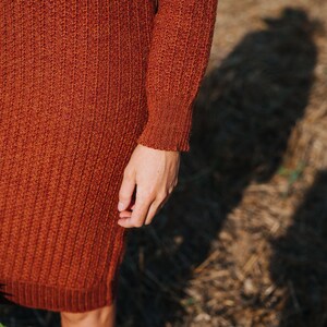 May include: A close-up of a rust-coloured knitted sweater dress with long sleeves. The dress is made of a soft, textured fabric and has a ribbed pattern.