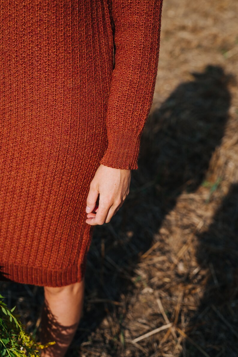 May include: A close-up of a rust-coloured knitted sweater dress with long sleeves. The dress is made of a soft, textured fabric and has a ribbed pattern.