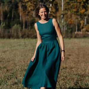 Peut inclure: Une robe sans manches de couleur teal avec un col rond et une jupe ample. La robe arrive au genou et semble &ecirc;tre en lin. La personne porte des chaussures noires et une montre noire. L'arri&egrave;re-plan pr&eacute;sente un champ et des arbres.