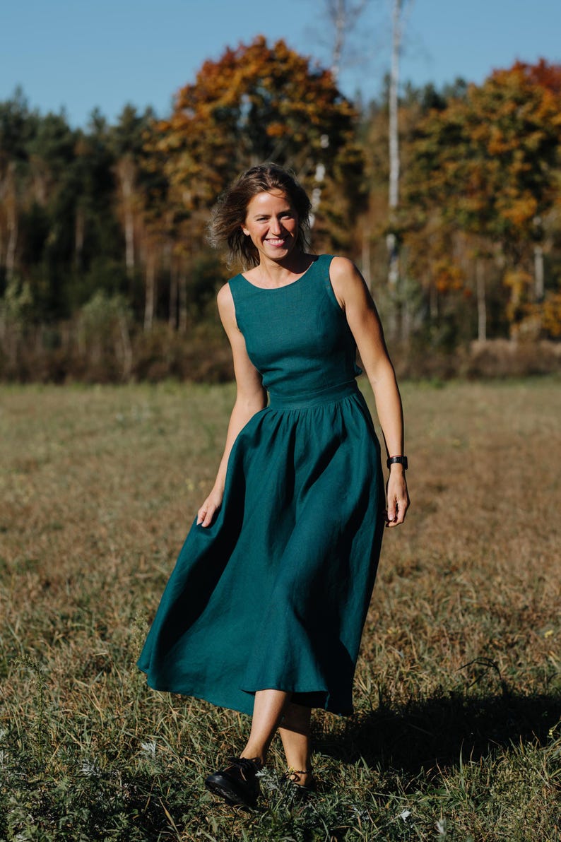 Peut inclure: Une robe sans manches de couleur teal avec un col rond et une jupe ample. La robe arrive au genou et semble &ecirc;tre en lin. La personne porte des chaussures noires et une montre noire. L'arri&egrave;re-plan pr&eacute;sente un champ et des arbres.