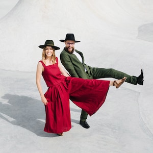 May include: A couple wearing hats and stylish clothing pose against a white background. The woman is wearing a red jumpsuit and the man is wearing a green suit.