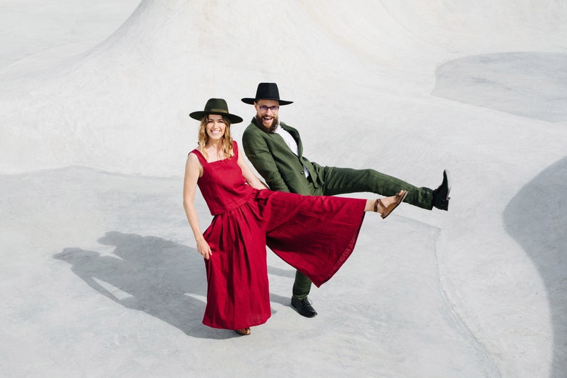 May include: A couple wearing hats and stylish clothing pose against a white background. The woman is wearing a red jumpsuit and the man is wearing a green suit.