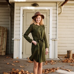 May include: A woman wearing a green linen dress with a brown hat and brown ankle boots stands on a wooden porch with fallen autumn leaves.