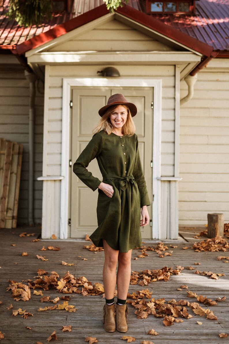 May include: A woman wearing a green linen dress with a brown hat and brown ankle boots stands on a wooden porch with fallen autumn leaves.