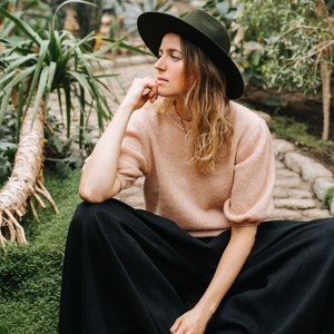 May include: A woman wearing a wide-brimmed black hat, a light pink sweater, and black wide-leg pants sits in a garden setting.