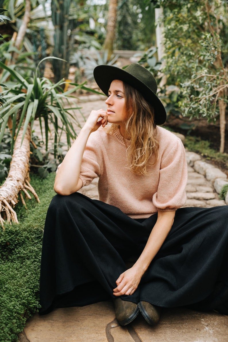 May include: A woman wearing a wide-brimmed black hat, a light pink sweater, and black wide-leg pants sits in a garden setting.