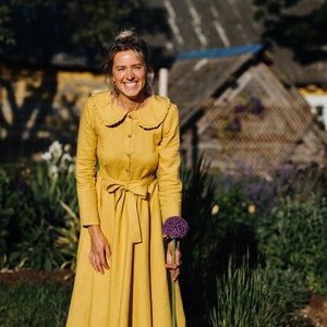 Pode incluir: Uma mulher em um vestido amarelo com gola de babados sorri enquanto segura uma flor roxa em um jardim.