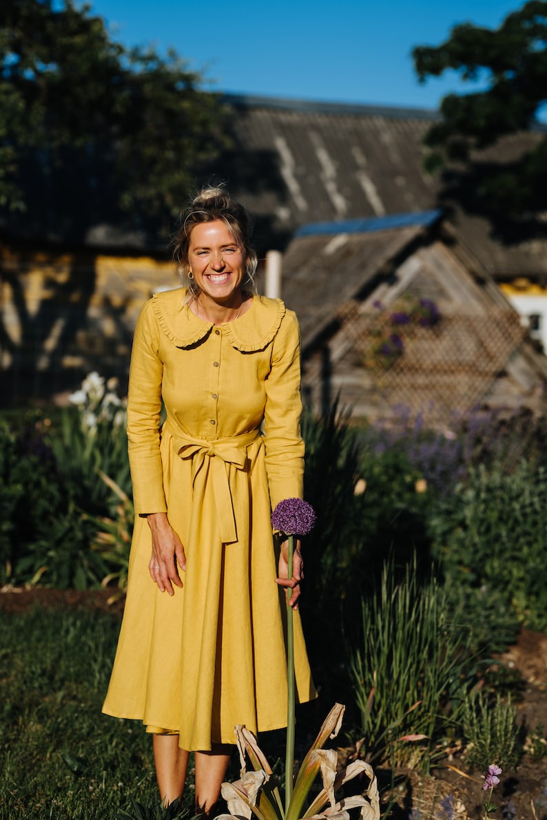Pode incluir: Uma mulher em um vestido amarelo com gola de babados sorri enquanto segura uma flor roxa em um jardim.