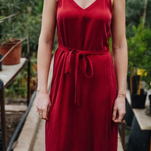 May include: A woman wearing a red sleeveless midi dress with a tie at the waist. The dress is made of a smooth, silky fabric.