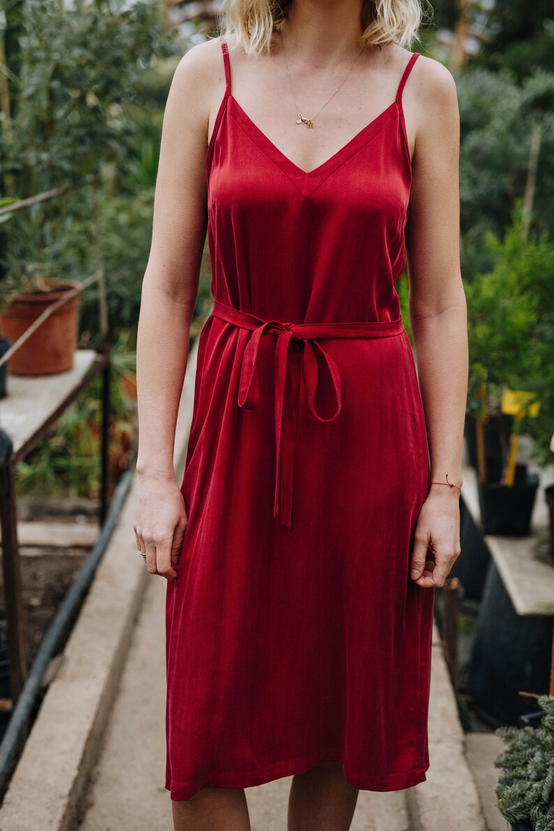 May include: A woman wearing a red sleeveless midi dress with a tie at the waist. The dress is made of a smooth, silky fabric.
