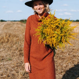 May include: A woman wearing a brown turtleneck sweater dress and a black fedora hat is holding a bouquet of yellow wildflowers in a field.