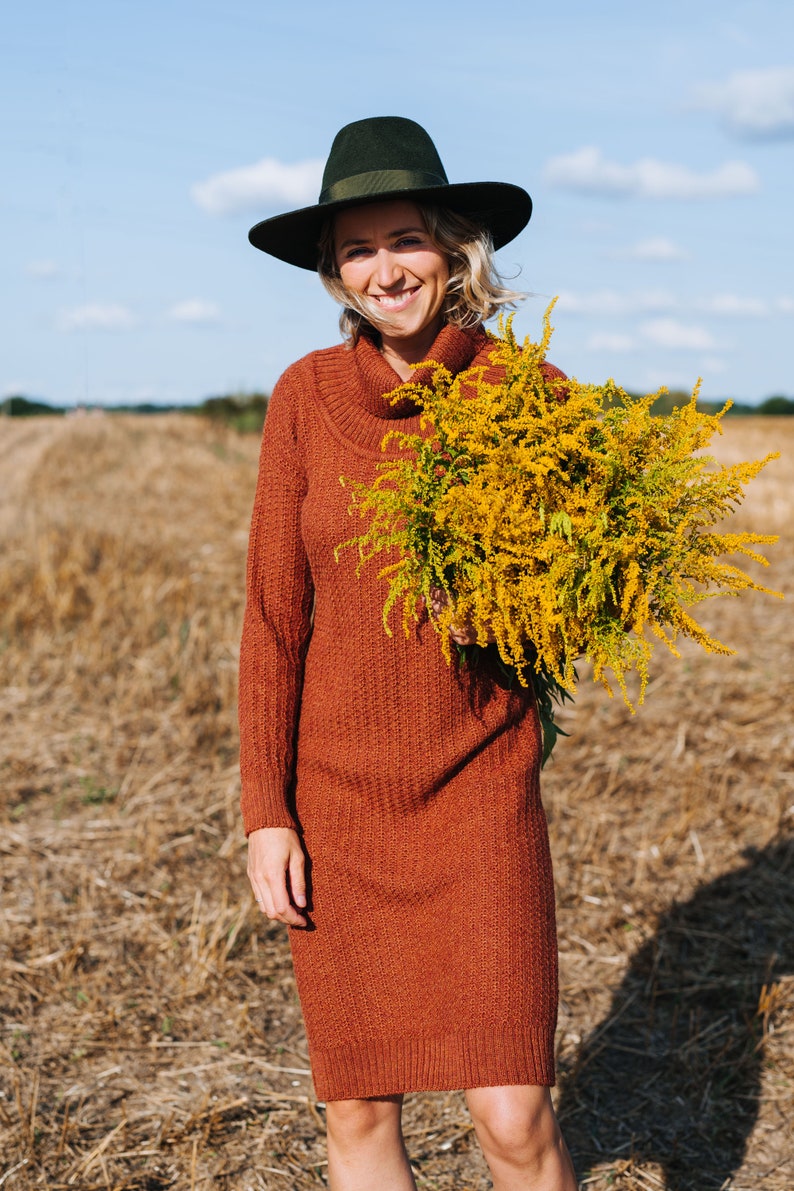 May include: A woman wearing a brown turtleneck sweater dress and a black fedora hat is holding a bouquet of yellow wildflowers in a field.