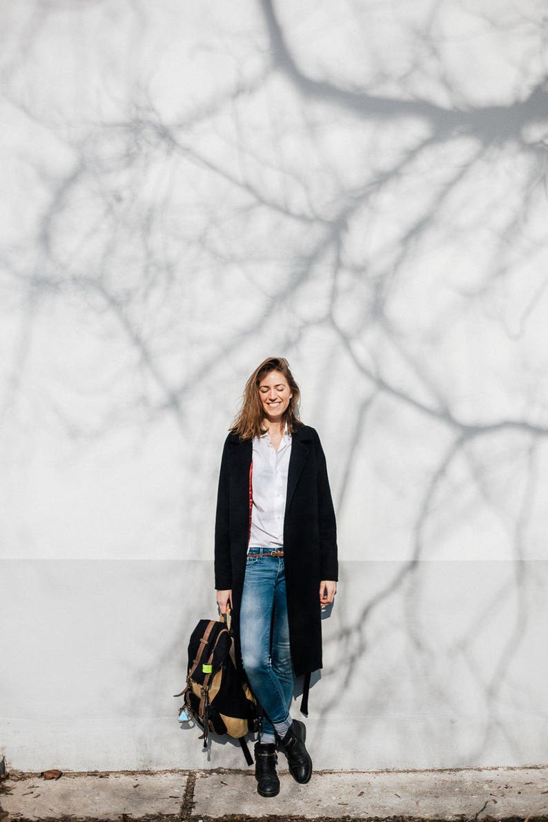 May include: A woman wearing a black coat, white shirt, and blue jeans stands in front of a white wall with a tree shadow. She is holding a black backpack.