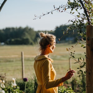 Pode incluir: Uma mulher com um vestido amarelo est&aacute; em um jardim, olhando para um campo. Ela est&aacute; com um coque e tem uma videira na m&atilde;o.