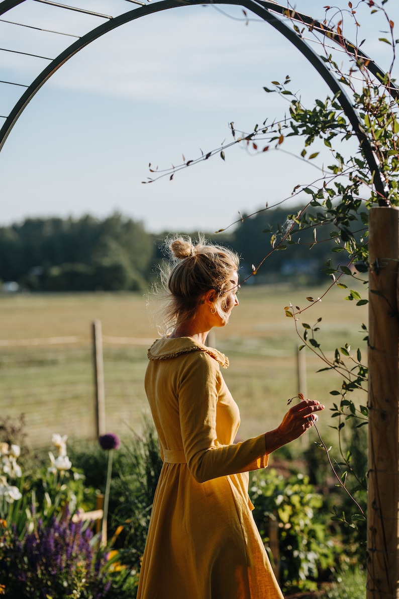 Pode incluir: Uma mulher com um vestido amarelo est&aacute; em um jardim, olhando para um campo. Ela est&aacute; com um coque e tem uma videira na m&atilde;o.