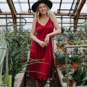 May include: A woman wearing a red dress and a green hat stands in a greenhouse surrounded by potted plants.