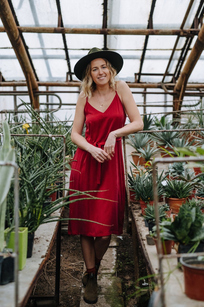 May include: A woman wearing a red dress and a green hat stands in a greenhouse surrounded by potted plants.
