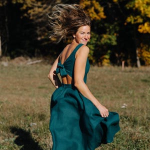 Peut inclure: Une robe de couleur teal avec un d&eacute;tail de n&oelig;ud dans le dos. La robe a une jupe fluide et un dos nu. La personne court dans un champ herbeux avec des arbres en arri&egrave;re-plan.