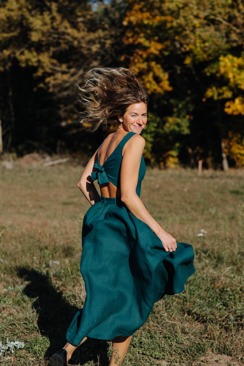 Peut inclure: Une robe de couleur teal avec un d&eacute;tail de n&oelig;ud dans le dos. La robe a une jupe fluide et un dos nu. La personne court dans un champ herbeux avec des arbres en arri&egrave;re-plan.