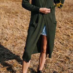 May include: A woman wearing a long, dark green cardigan with a white tank top underneath. She is standing in a field of dry grass and holding a small bouquet of yellow flowers.