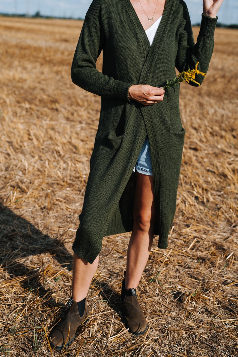 May include: A woman wearing a long, dark green cardigan with a white tank top underneath. She is standing in a field of dry grass and holding a small bouquet of yellow flowers.