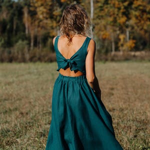 Peut inclure: Une robe de couleur teal avec un n&oelig;ud dans le dos. La robe a un haut sans manches et une jupe fluide. La personne se tient dans un champ avec des arbres en arri&egrave;re-plan. La robe est en tissu doux et fluide.