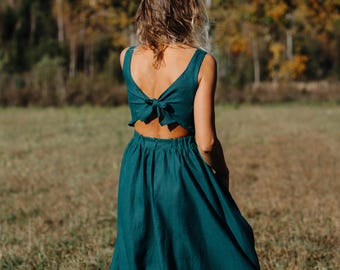 May include: A teal-colored dress with a bow detail on the back. The dress has a sleeveless top and a flowing skirt. The person is standing in a field with trees in the background. The dress is made of a soft, flowing fabric.