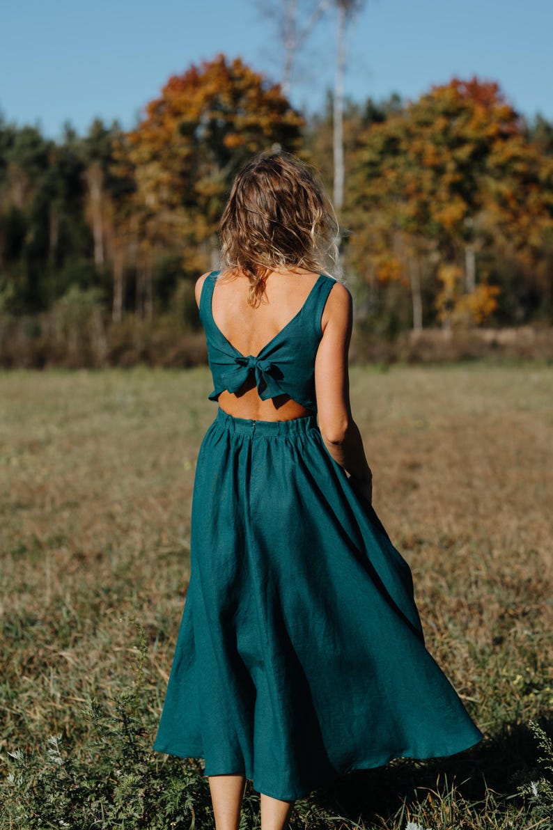 Peut inclure: Une robe de couleur teal avec un n&oelig;ud dans le dos. La robe a un haut sans manches et une jupe fluide. La personne se tient dans un champ avec des arbres en arri&egrave;re-plan. La robe est en tissu doux et fluide.