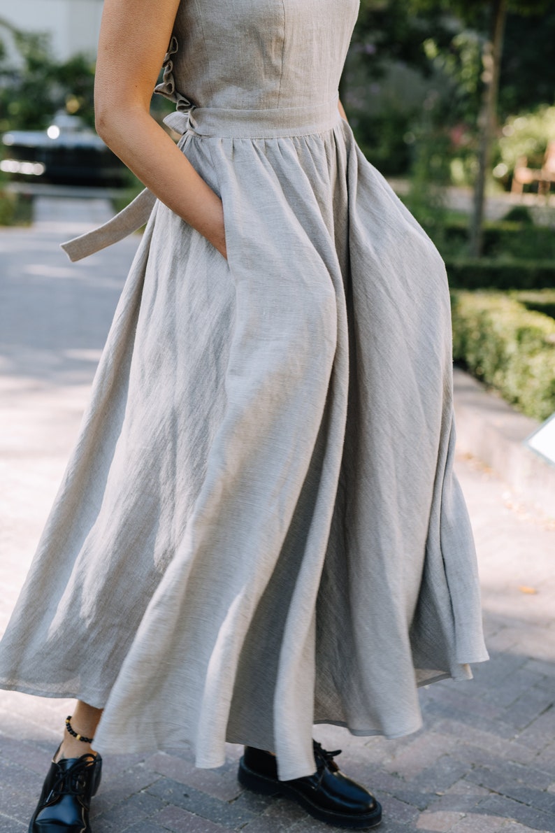 May include: A light gray linen dress with a gathered skirt and a tie at the waist. The dress has a simple, elegant design and is perfect for a summer day.