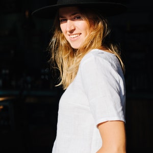 Op de afbeelding: Een vrouw draagt een wit linnen shirt en een zwarte fedora hoed. Ze glimlacht en kijkt naar de camera.