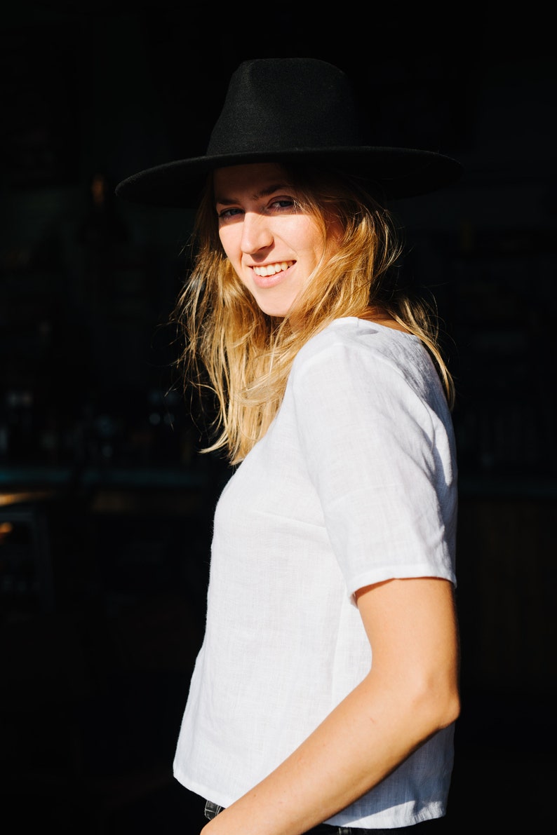 Op de afbeelding: Een vrouw draagt een wit linnen shirt en een zwarte fedora hoed. Ze glimlacht en kijkt naar de camera.