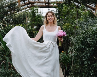 Pode incluir: Uma mulher com um vestido branco de linho segura um buquê de peônias cor-de-rosa em uma estufa. O vestido tem um design simples com um laço na cintura. A mulher está sorrindo e olhando para a câmera.
