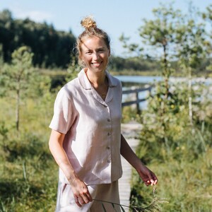 May include: A light pink linen pajama set, including a button-down shirt and shorts. The short-sleeved shirt has a collar and buttons down the front. The shorts have a relaxed fit. The set is worn outdoors on a wooden walkway.