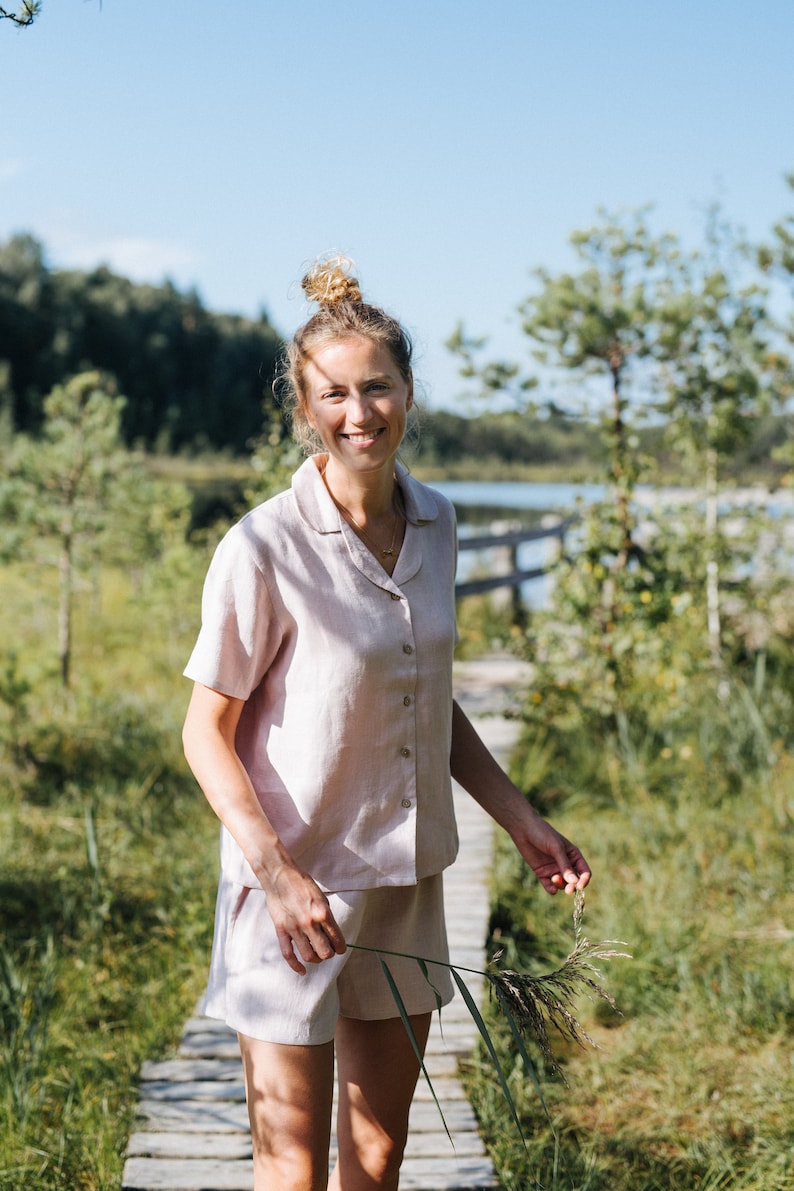 May include: A light pink linen pajama set, including a button-down shirt and shorts. The short-sleeved shirt has a collar and buttons down the front. The shorts have a relaxed fit. The set is worn outdoors on a wooden walkway.