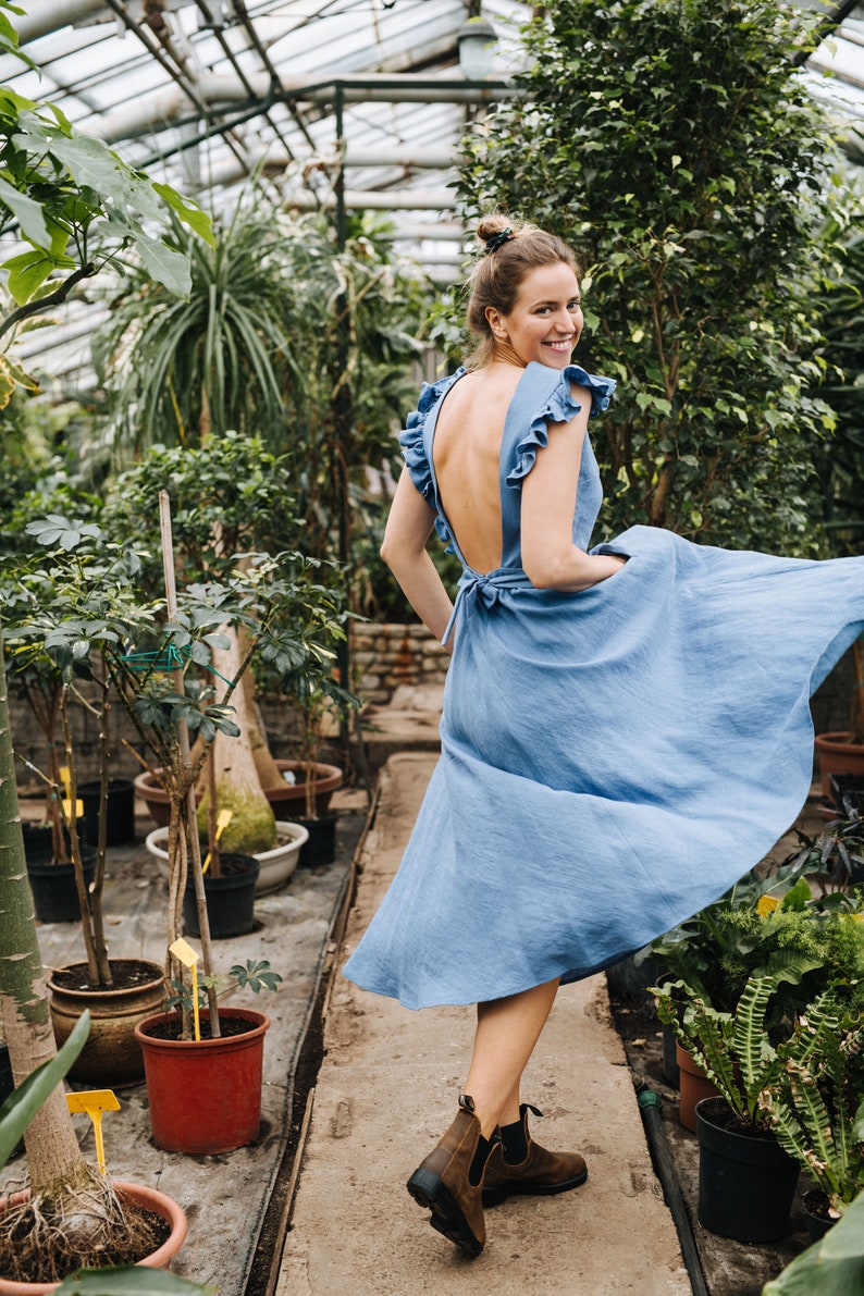 Puede incluir: Una mujer con un vestido azul claro con la espalda descubierta y volantes en las mangas est&aacute; de pie en un invernadero. Est&aacute; sonriendo y mirando por encima del hombro. El vestido ondea con la brisa.