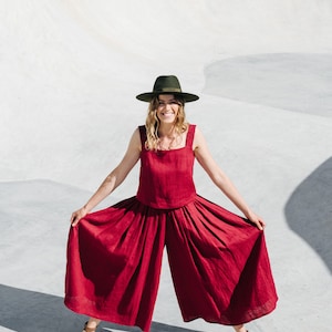 May include: A woman wearing a red linen jumpsuit with wide legs and a green hat. She is standing on a concrete surface and smiling at the camera.