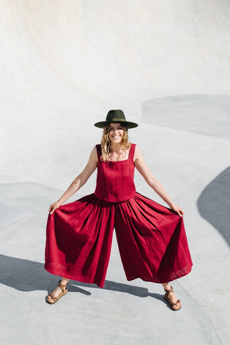 May include: A woman wearing a red linen jumpsuit with wide legs and a green hat. She is standing on a concrete surface and smiling at the camera.