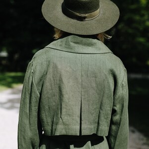 May include: A person wearing a green linen trench coat with a wide brimmed green felt hat.