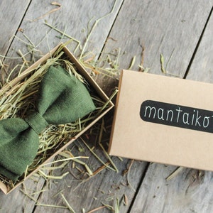 K&ouml;nnte beinhalten: Ein dunkelgr&uuml;ner Fliege aus Leinen in einer braunen Schachtel mit zerkn&uuml;lltem Papier. Die Schachtel hat ein schwarzes Etikett mit dem Text "mantai kotai".