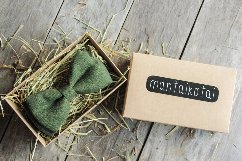 K&ouml;nnte beinhalten: Ein dunkelgr&uuml;ner Fliege aus Leinen in einer braunen Schachtel mit zerkn&uuml;lltem Papier. Die Schachtel hat ein schwarzes Etikett mit dem Text "mantai kotai".