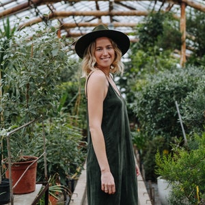 Puede incluir: Una mujer con un vestido verde oscuro y un sombrero de ala ancha se encuentra en un invernadero. Sonríe y mira a la cámara. El invernadero está lleno de plantas y tiene una estructura de metal.