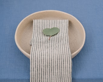 May include: A beige ceramic bowl holds a folded striped napkin with a green leaf on a blue tablecloth. The napkin has vertical stripes in a muted green and white. The leaf is heart-shaped.