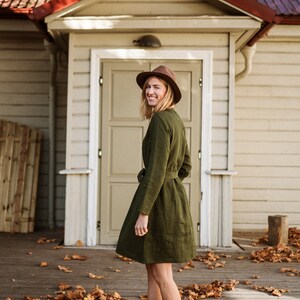 May include: A woman wearing a brown hat and a green linen dress walks on a wooden deck covered in autumn leaves.