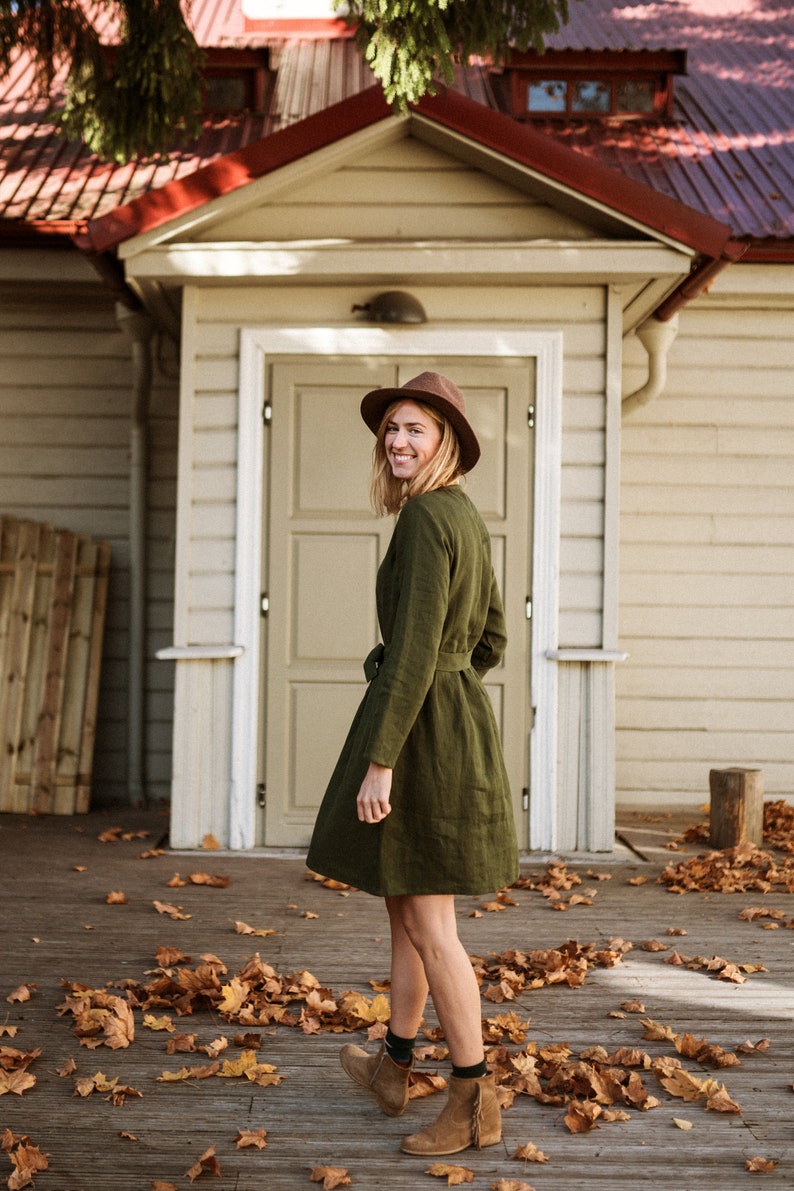 May include: A woman wearing a brown hat and a green linen dress walks on a wooden deck covered in autumn leaves.